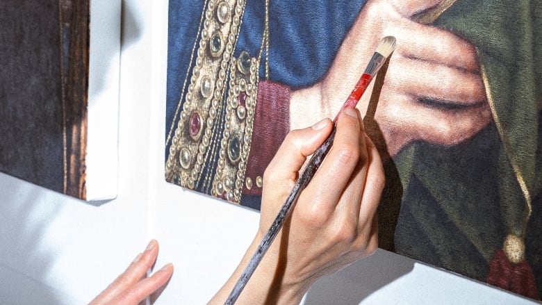 A hand holding a brush is seen painting a hand on a canvas that's hanging on the wall. 