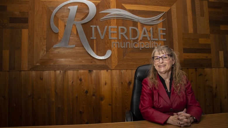 A woman sits at a desk with a Riverdale sign behind her.