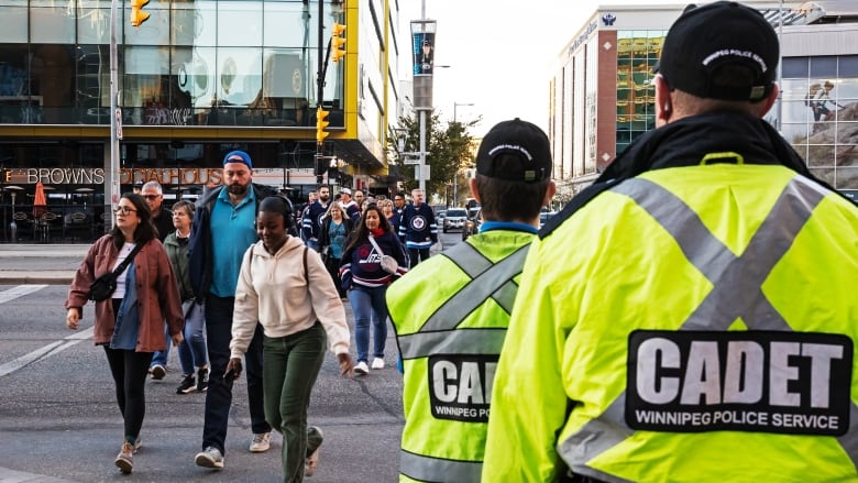 Two people wearing jackets labelled 'Cadet Winnipeg Police Service' look on as people wearing Winnipeg Jets jerseys cross the street towards them.