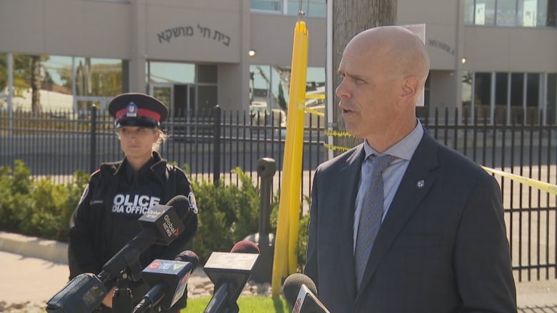 A man in a suit stands in front of microphones on a city street outside a school where police tape is up in the yard. A female officer stands off to his side.