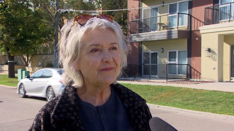 A woman in a sweater stands outside an apartment block, listening as a microphone is held up in front of her.