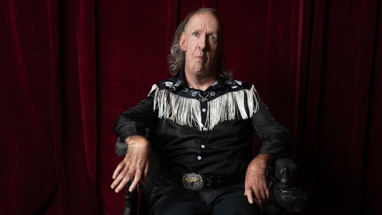 A man in a fringed western shirt sits in a wheelchair in front of a deep red curtain.