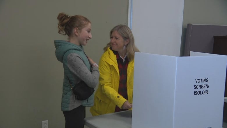 A woman with blonde hair and wearing a yellow rain jacket stands next to a table that has white cardboard standing up with the text Voting Screen written across it. A young girl stands next to her.