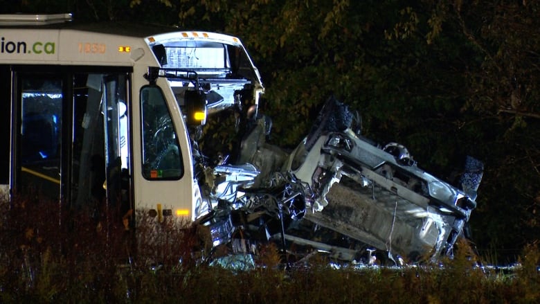 On a rural stretch of highway, at night, a transit bus with its front end smashed in rests against a totaled pickup truck which has rolled over.