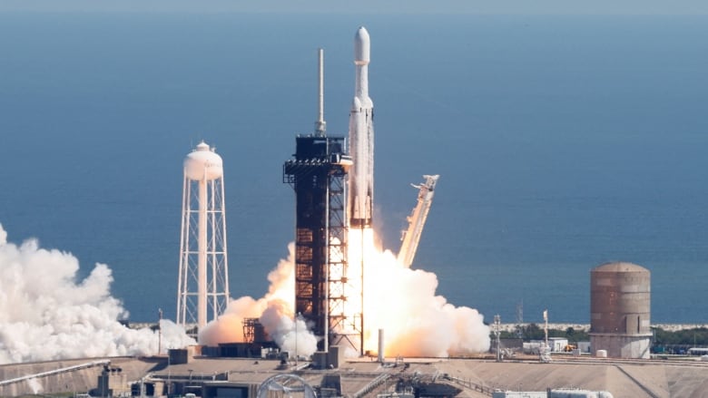 A white rocket takes off from the scaffolding that previously held it, with a large plume of flame and smoke underneath it.  