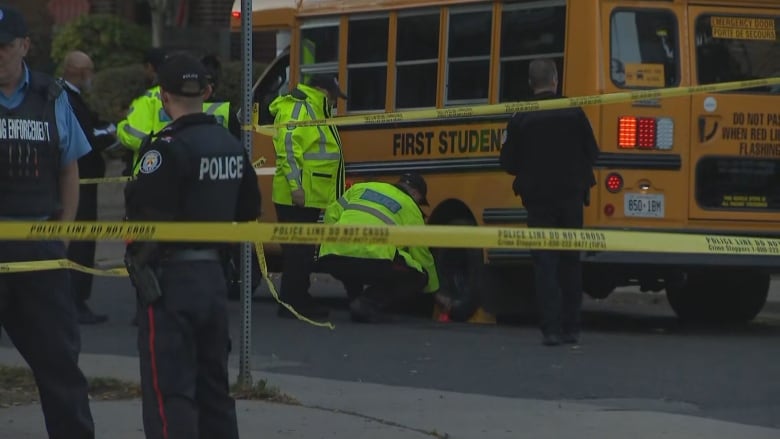 Emergency responders examine a school bus parked on the side of a city street on a late fall afternoon. There is police tape surrounding the scene.