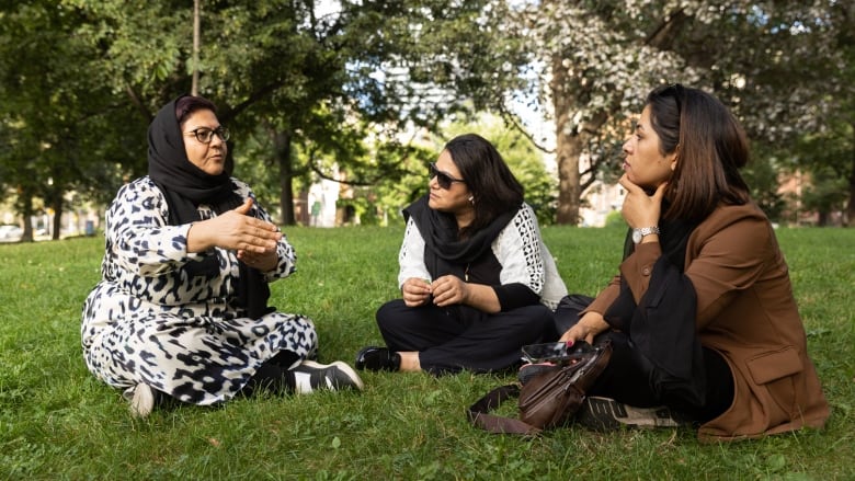 Three women sit on a grassy green hill with trees in the background