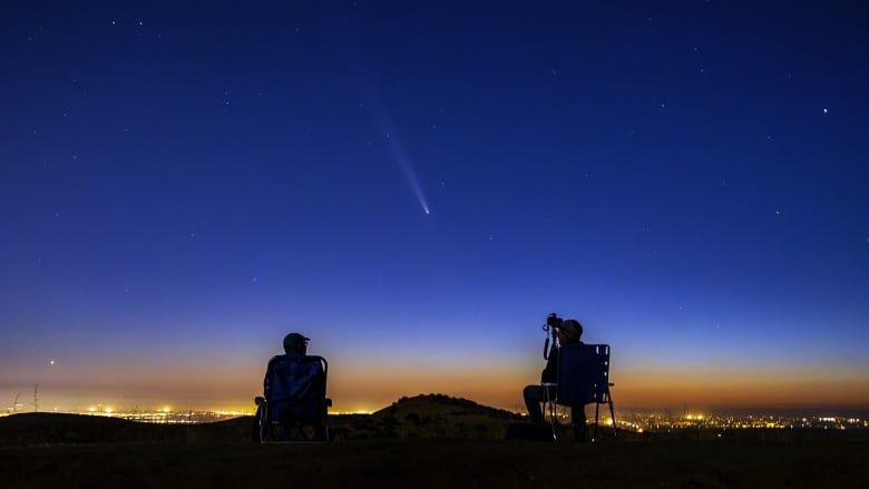 Comet in the night sky between two people in silhouette, sitting in lawn chairs watching and taking photos.