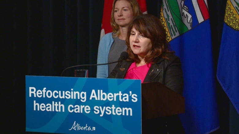 two women stand in front of a row of flags. one woman stands in front of a podium and speaks into a microphone.