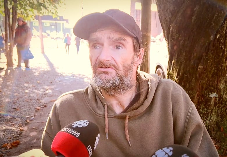 A man in a grey hoodie wearing a bluish grey ballcap with a salt-and-pepper beard and moustache speaks on a sidewalk.