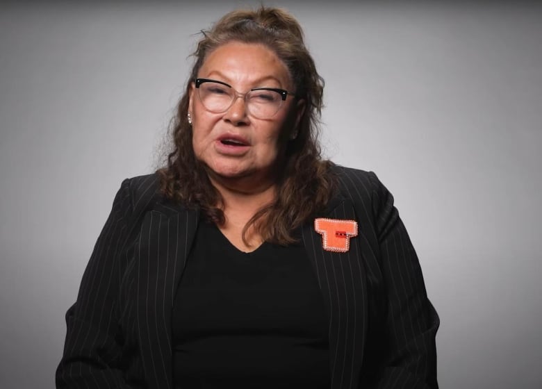 A woman stands in front of a grey and white background. She is wearing a black suit with a black shirt. Pinned to her left lapel is a orange shirt pin.