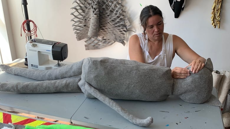 Women sews large doll at table.