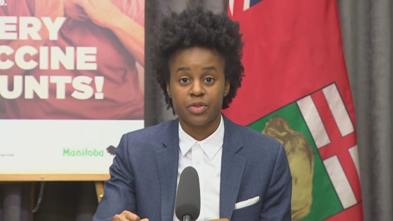 A person with black hair, and wearing a grey suit, sits at a desk speaking into a microphone.