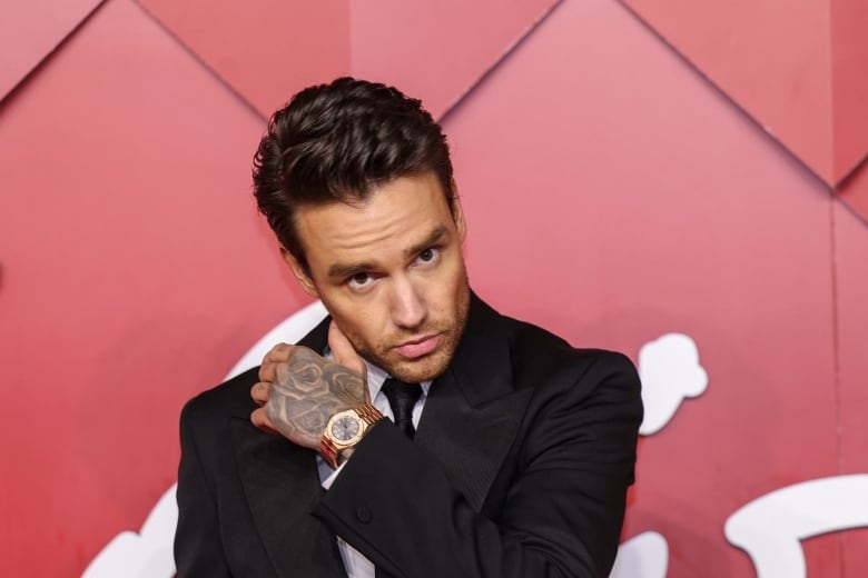 A dark haired man in a suit raises his tattooed hand in a red carpet photo.