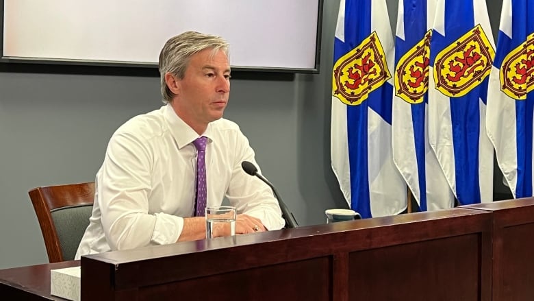 Premier Tim Houston is seen seated in front of a microphone with Nova Scotia flags behind him.