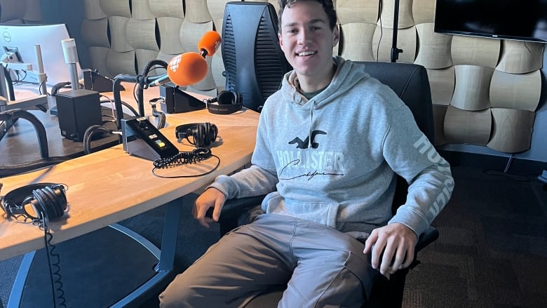 A young man in a grey hoodie sits in front of a table in a radio studio.