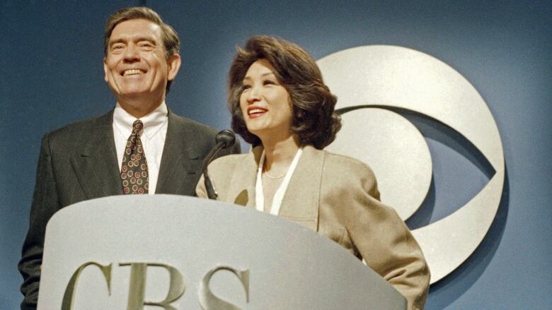 A man and a woman both wearing suits stand smiling behind a podium that says 'CBS.'