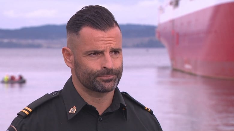 A fisheries officer in uniform stands in front of a red ship in the inner harbour in Victoria.