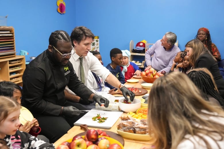 Prime Minister Justin Trudeau cuts fruit next to chef Jason Simpson at photo opportunity as they prepare food for a lunch program at the Boys and Girls Club East Scarborough, in Toronto, before an announcement to launch a National School Food Program on April 1. Liberal MP John MacKay, Finance Minister Chrystia Freeland and Social Development Minister Jenna Sudds are also seated around the low table with local kids of diverse backgrounds.