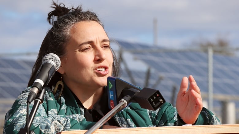 A woman with a green jacket stands in front of a podium. Behind her solar panels can be seen. 