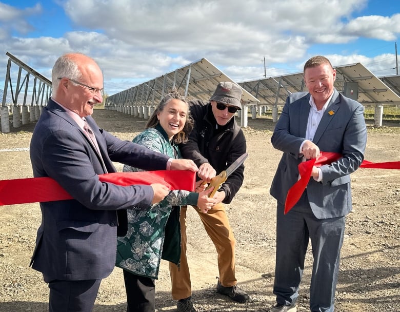 Three men and a woman cut a red ribbon. A field of solar panels can be seen behind them.