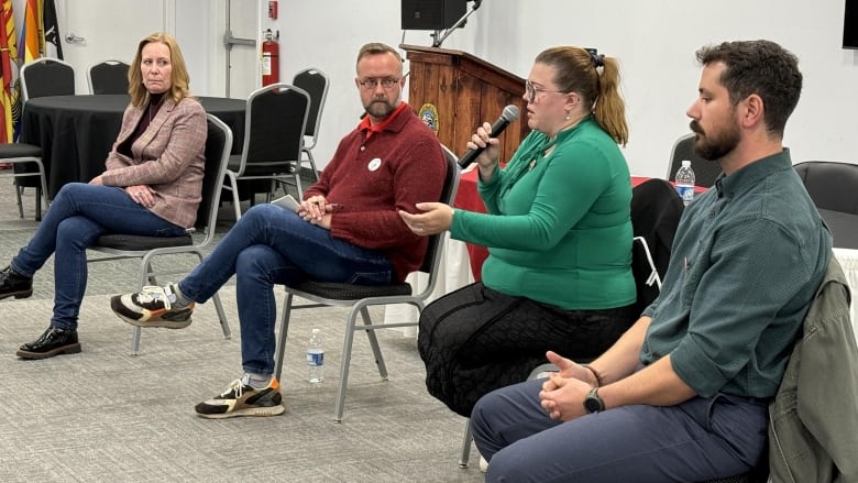 Four people sit on chairs in a line inside a room with white walls. On one chair, a woman wearing green speaks into a microphone.