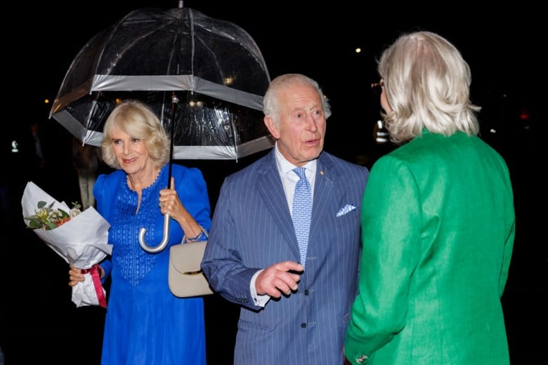 Two people talk while another person with an umbrella stands behind.