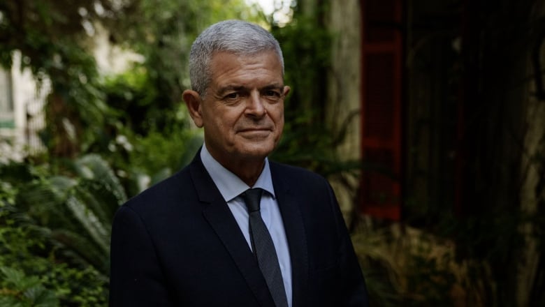 A man with grey hair in a dark suit poses for a photo. There is a lush garden behind him. 