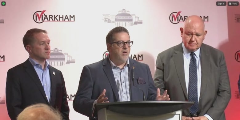 A man wearing a suit jacket stands at a podium between two other men in suits. He's wearing glasses and is speaking with his hands, as both are raised for emphasis.