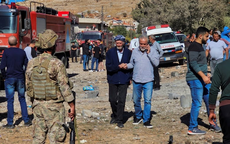 A soldier stands guards as people inspect the site of a rocket attack.