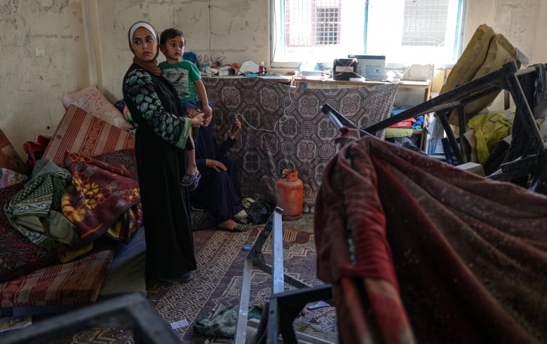 An adult carries a child inside a room where the furniture is disarranged.