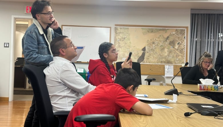 A man in a shirt and tie with children on either side of him at a board room table. A man speaking on the phone stands behind him. 