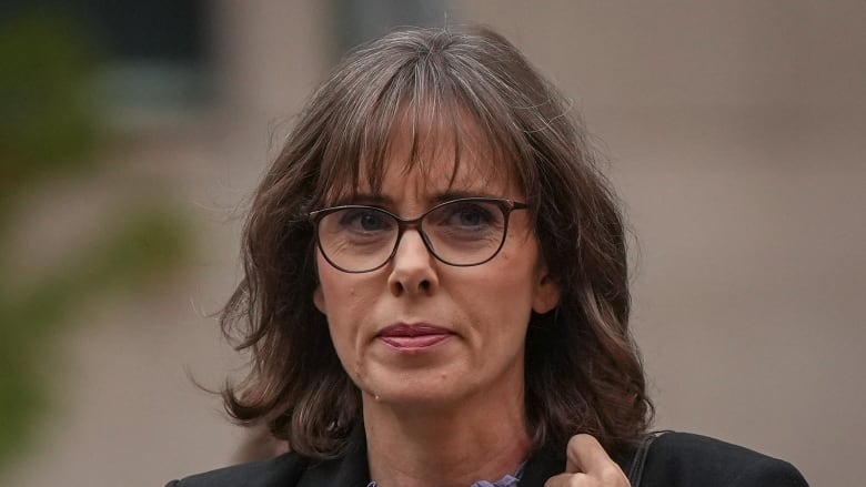 A woman in a purple shirt and a black blazer is seen wearing glasses as she walks into a building.