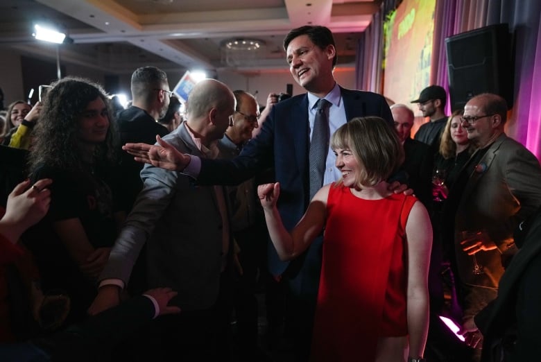 A man in a suit greets supporters alongside his wife, who is wearing a red dress.