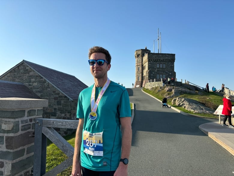 A man stands in front of Cabot Tower. He is wearing a green tee shirt and a medal around his neck.