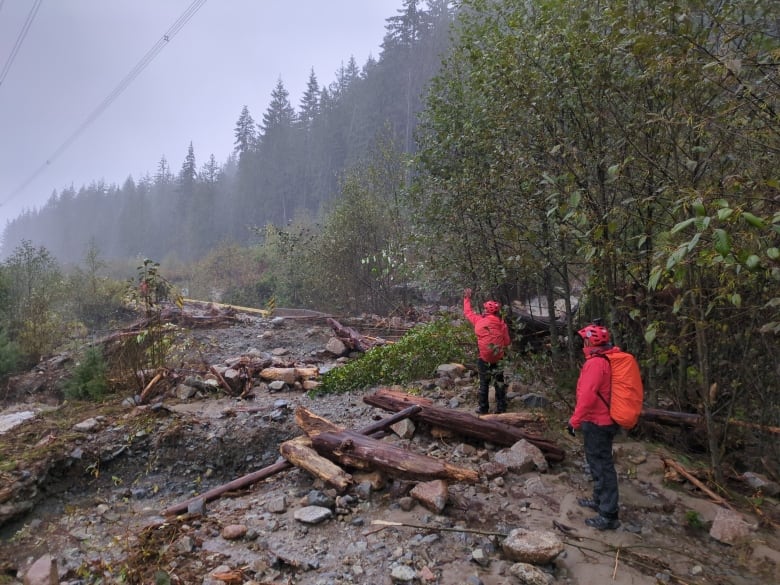 Two people dressed in red jackets and helmets wave to unseen people across a mudslide.