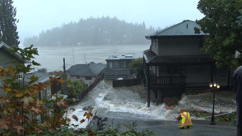 Scenes from a flood shows a man in reflector jacket crouched down in a road that slopes towards homes inundated by flood. 