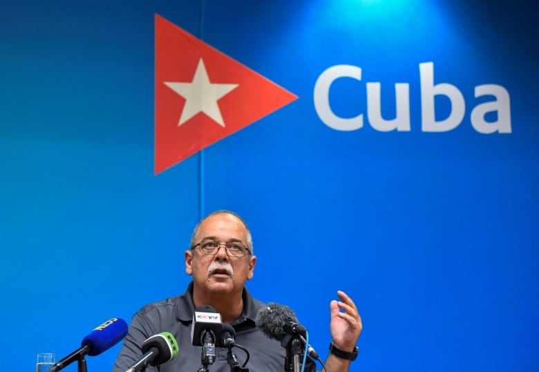 A person sitting in front of a wall featuring the Cuban flag speaks into microphones during a news conference.