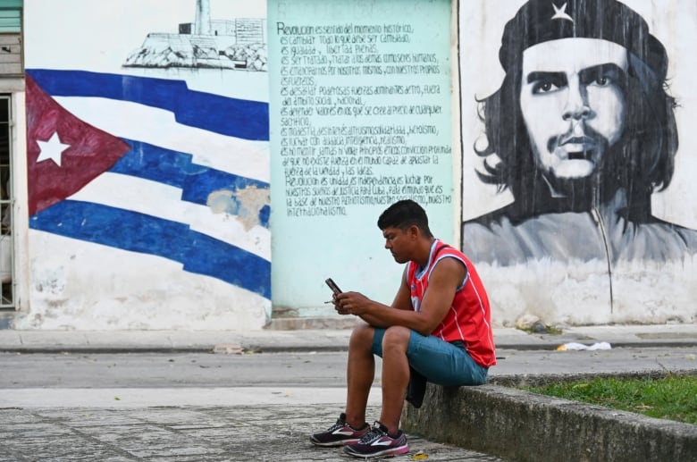 A person uses their mobile phone while sitting on a curb as a mural of depicting the Cuban flag and Che Guevara is seen in the background.