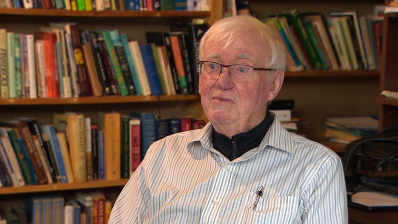 A man in a white shirt sitting in front of a bookshelf. 