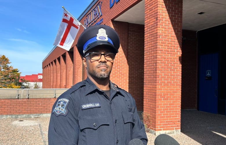 A uniformed police officer stands in front of a brick building. There are two mics in front of him. 