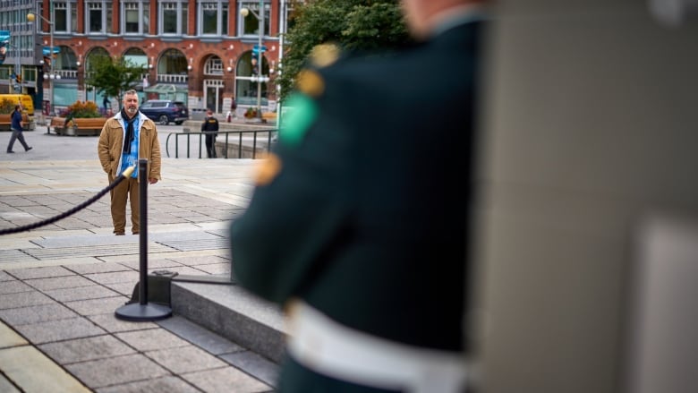 A man in a brown jacket looks at the National War Memorial, where the shoulder of a soldier standing sentry is visible in the foreground, out of focus.