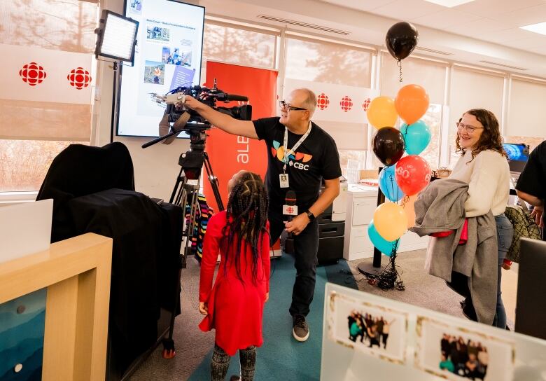 a man shows a young girl how a tv camera and prompter works, as a woman looks on