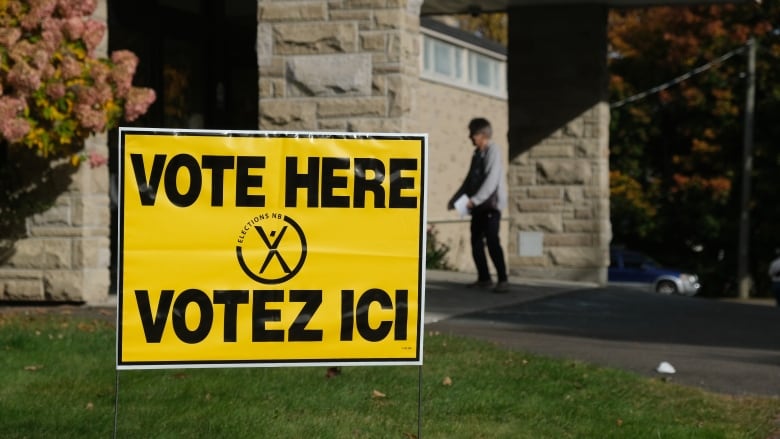 A yellow and black 'Vote here' Elections NB sign in the foreground and a person entering a door holding a piece of paper in the background.