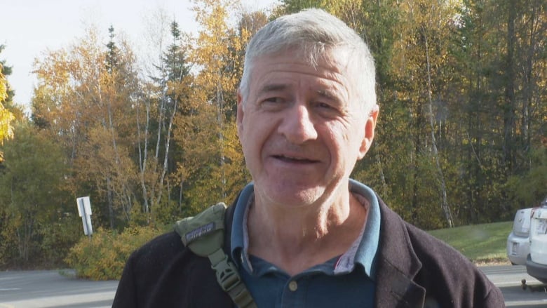 A man with short grey hair, in a parking lot, with trees behind him.