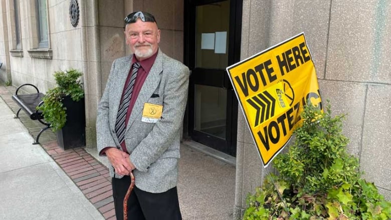 A man with grey hair and goatee, wearing a grey blazer, grey and black diagonally striped tie, and burgundy dress shirt, holding a cane, outside a polling station.