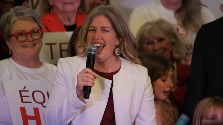 Smiling woman holds a microphone, with crowd behind her. 