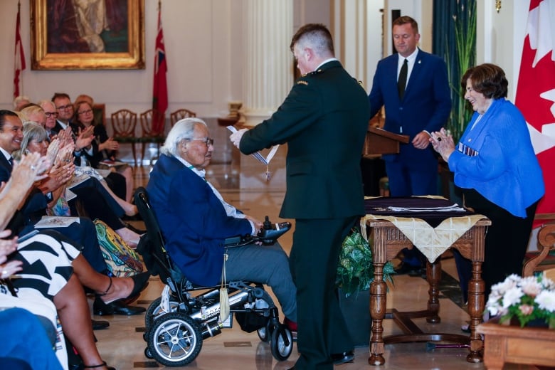 Murray Sinclair in a wheelchair receiving Manitoba's highest honour, during a ceremony at the provincial legislature with people clapping in the audience behind him.