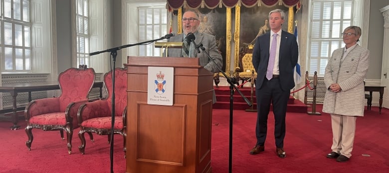 A man in a suit speaks at a podium as a man and a woman in suits look on. The room in Nova Scotia's Province House has a red carpet and two ornate red chairs. 