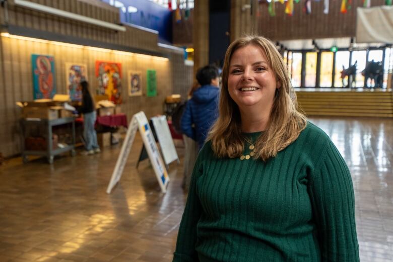 A person wearing a pine green sweater stands inside a building. They are smiling.